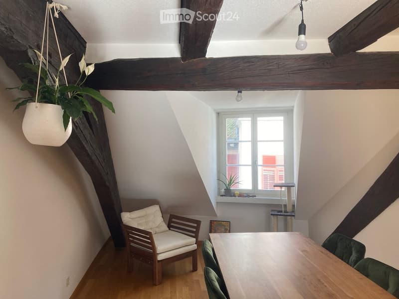 Cozy attic room with wooden beams, slanted ceilings, and large window. Features a wooden table with green chairs, a white cushioned chair, and a hanging plant.
