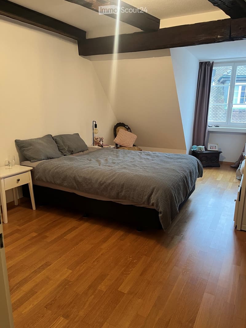 Cozy attic bedroom with wooden flooring, slanted ceilings, and large window offering a view of the roof. Features a double bed with grey bedding, nightstands, a chair, and decorative items.