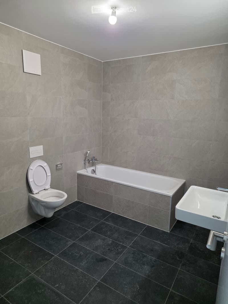 Modern bathroom with grey tiled walls, black tiled floor, toilet, bathtub, and sink.