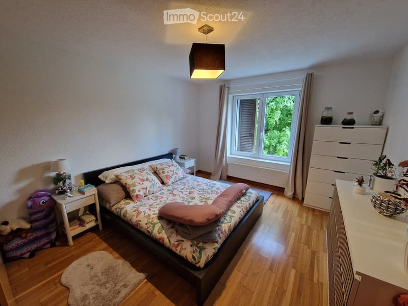 Cozy bedroom with a large bed, nightstand, lamp, and a dresser with a mirror. The room features wooden flooring and a large window with curtains.