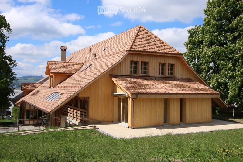 A two-story wooden house with a red-tiled roof, multiple windows, and a raised ground floor entrance. The house is surrounded by a well-maintained lawn and trees.