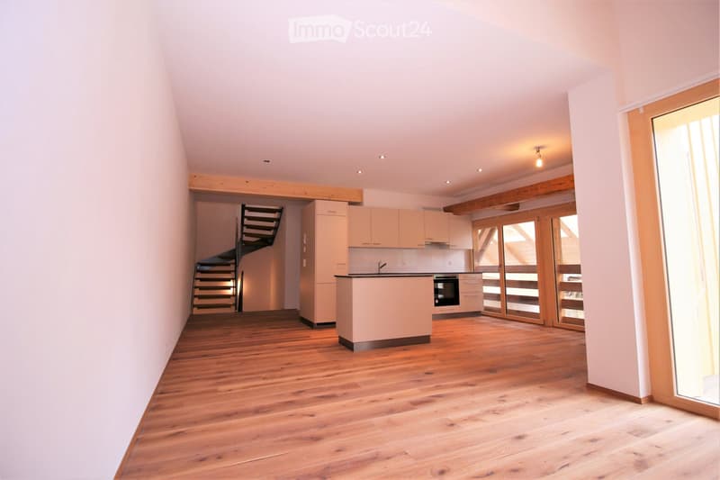 Open-plan kitchen and living area with light wooden flooring, white cabinetry, and large glass doors leading to a balcony. The space is empty, with no furniture or appliances visible.