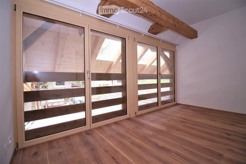 Empty room with wooden flooring, white walls, and large sliding glass doors with wooden frames and blinds. Exposed wooden beams on the ceiling.