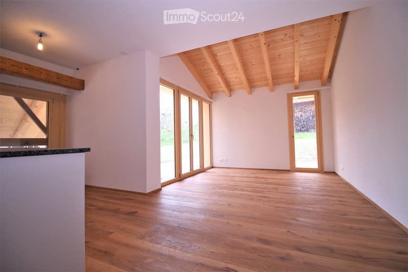 Empty room with wooden flooring, white walls, exposed wooden beams on the ceiling, and large sliding glass doors leading outside.