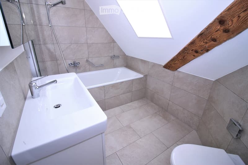 Modern bathroom with a large sink, bathtub, toilet, and a skylight window. Features include a wooden slanted ceiling beam and tiled walls and floor.