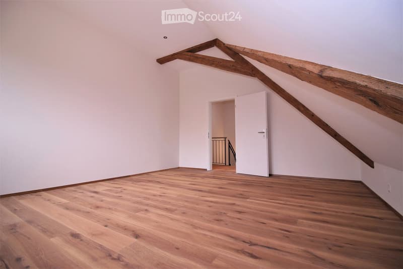 Empty attic room with wooden floors, white walls, and exposed wooden beams. A white door leads to a staircase area.