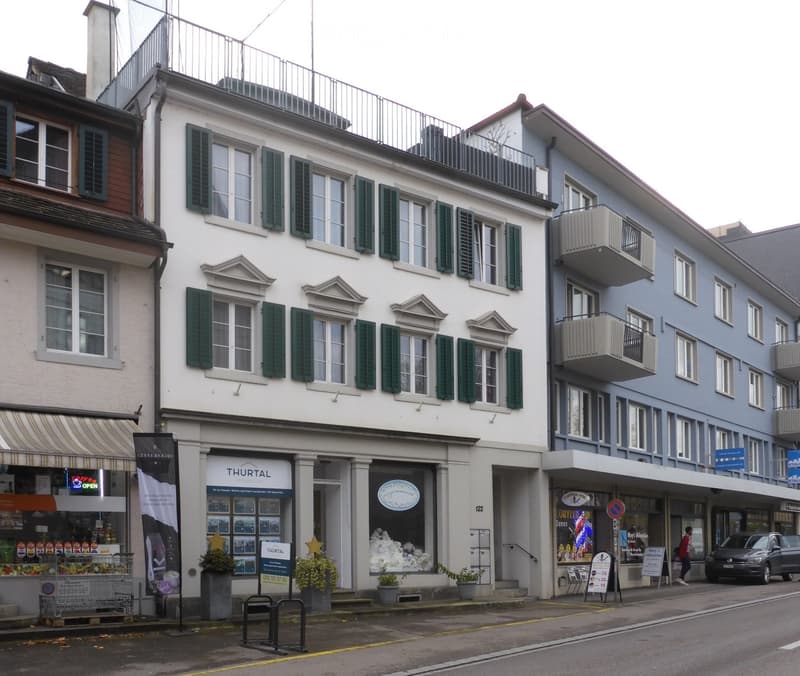 A multi-story building with white facade, green shutters, and multiple windows. The ground floor features commercial storefronts with signage, including 'THURTAL' and 'COFFEE DAMEN'. There are potted plants near the entrance and a small awning over one of