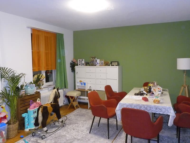 A cozy living room with a mix of furniture and toys, including a rocking toy horse, a small table with chairs, a cabinet with framed pictures, and a potted plant near the window.