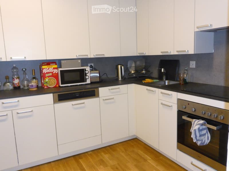 Modern kitchen with white cabinets, stainless steel appliances, and a black countertop. Visible items include a microwave, oven, toaster, sink, and various bottles and cups on the counter.