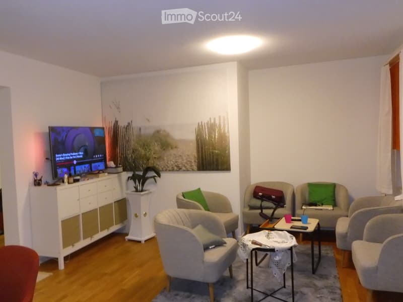 Modern living room with wooden flooring, multiple seating options including sofas and chairs, a television, a small table with cups, and a potted plant near a large picture frame.