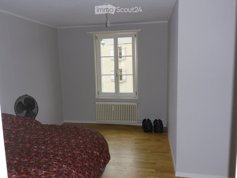 Bedroom with wooden floor, bed with patterned bedspread, fan, radiator, and two headphones on the floor.