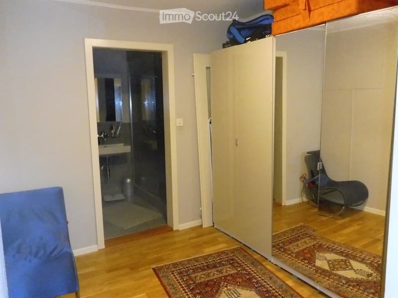 A furnished room with light wooden flooring, a large wardrobe, a blue chair, a patterned rug, and an open door leading to a bathroom.