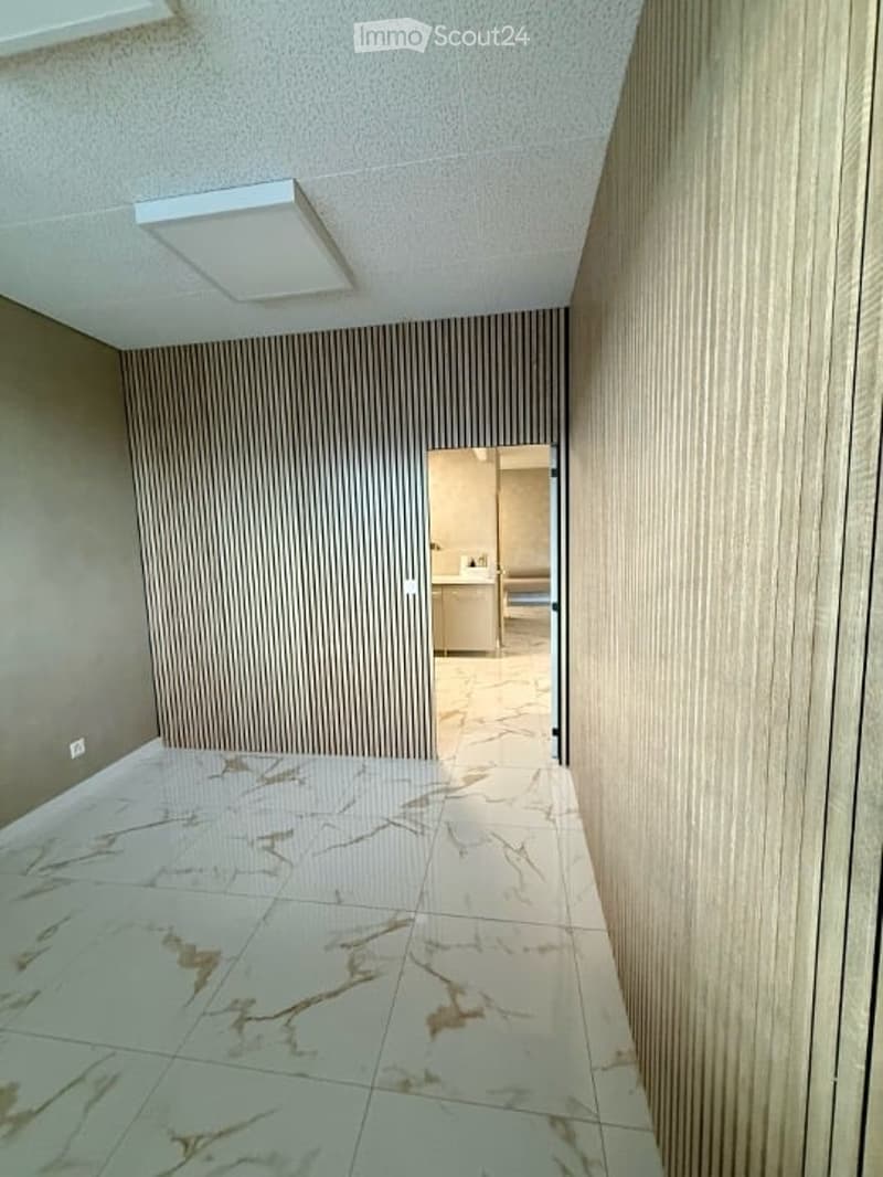 A clean, modern hallway with light-colored walls, a marble tiled floor, and a recessed ceiling light. An open doorway leads to a small, furnished room visible in the background.