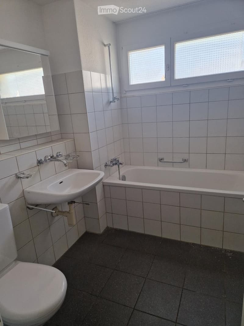 Bathroom with white tiled walls and floor, featuring a sink, bathtub, toilet, and mirror. Windows provide natural light.