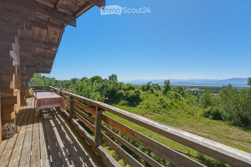 Wooden balcony with a table and chair, overlooking a lush green valley and distant mountains under a clear blue sky.