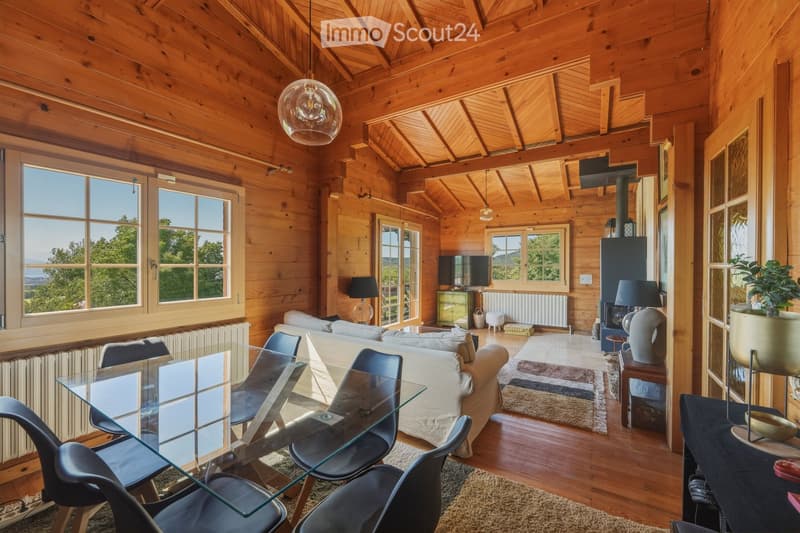 Cozy living room with exposed wooden beams, large windows offering a scenic view, a white sofa, a glass dining table with modern chairs, a fireplace, and decorative lamps.