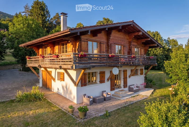 A two-story wooden house with a large balcony, surrounded by greenery and a well-maintained lawn. The house features a mix of wooden and white exterior walls, multiple windows with shutters, and a chimney. Outdoor seating includes a sofa, chairs, and a sm