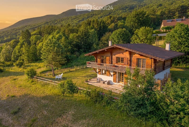 A wooden house with a large balcony, surrounded by greenery and a scenic mountain backdrop.