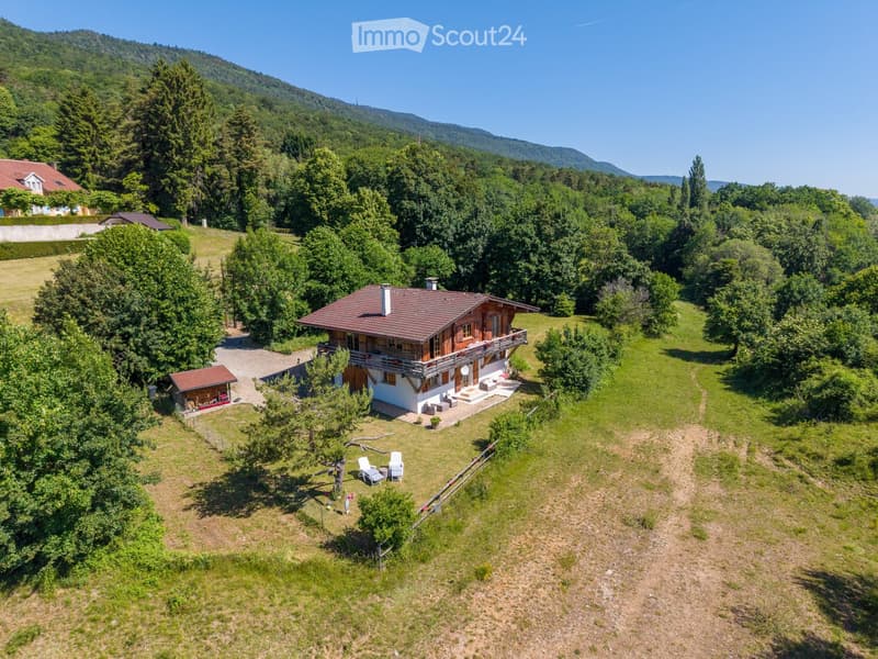 A two-story wooden house with a red-tiled roof, surrounded by a lush green landscape with trees and a small garden area. The house features a balcony and outdoor seating area.