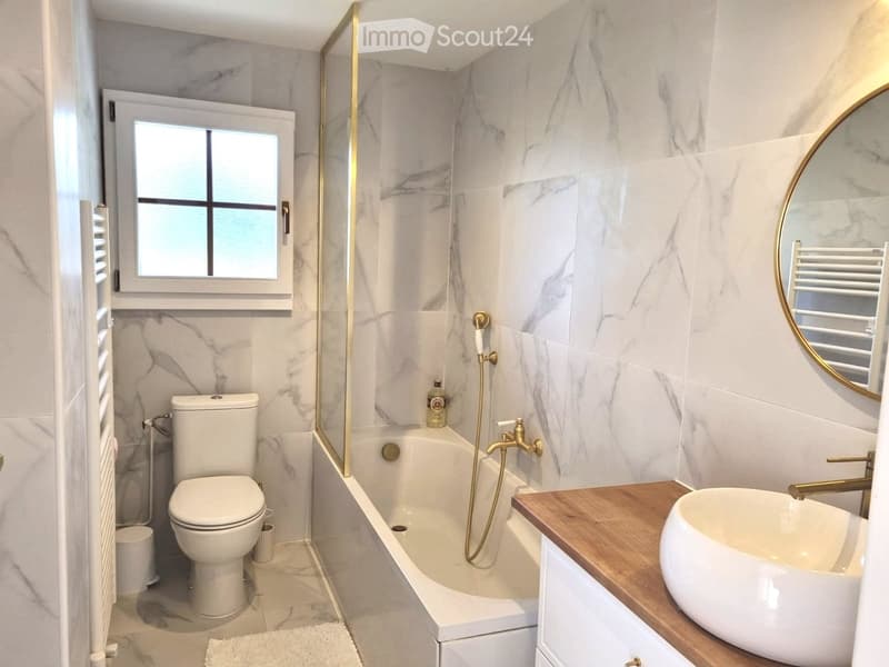 Modern bathroom with marble tiles, a bathtub with a gold showerhead, a toilet, a sink with a wooden countertop, and a large round mirror.