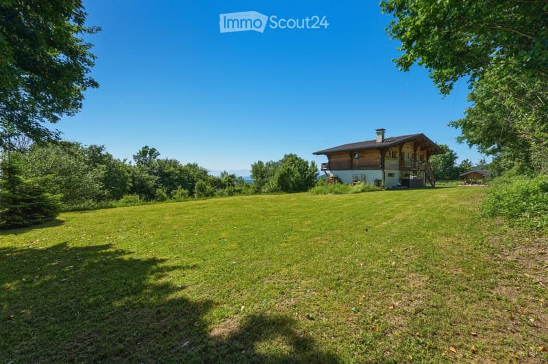 A large, well-maintained lawn with a wooden house featuring a balcony and staircase, surrounded by trees and greenery.