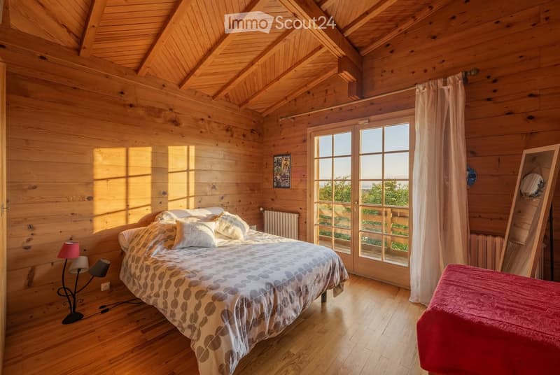 Cozy wooden bedroom with slanted wooden ceiling, large window with white curtains, bed with patterned bedspread and pillows, bedside lamp, mirror, radiator, and a picture frame on the wall.