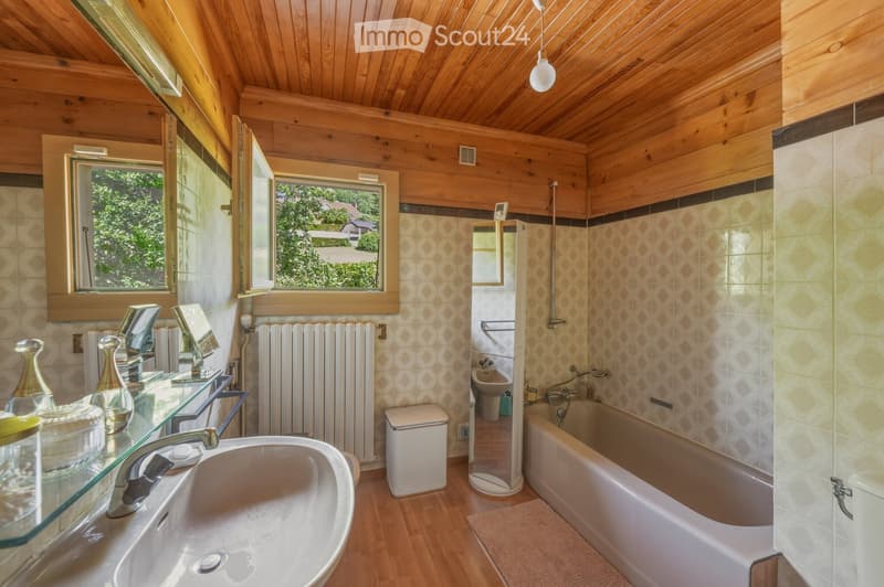 Bathroom with wooden ceiling and walls, featuring a sink, bathtub, and radiator. The room has large windows with wooden frames and a view of greenery outside.