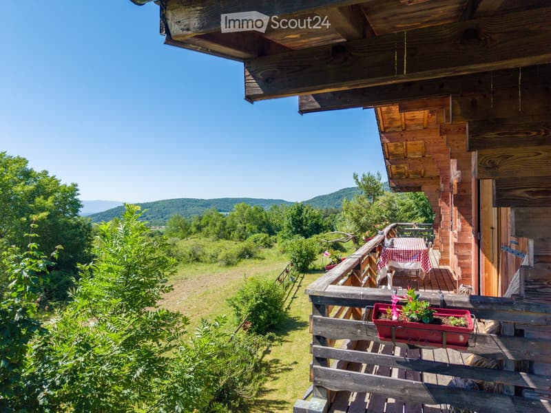 Wooden balcony with a table, chairs, and flower pots, overlooking a green landscape with hills in the background.