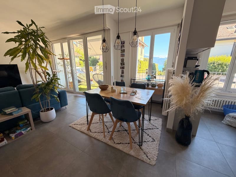 Bright living and dining area with large windows and a view, featuring a sofa, dining table with chairs, pendant lights, and decorative plants.