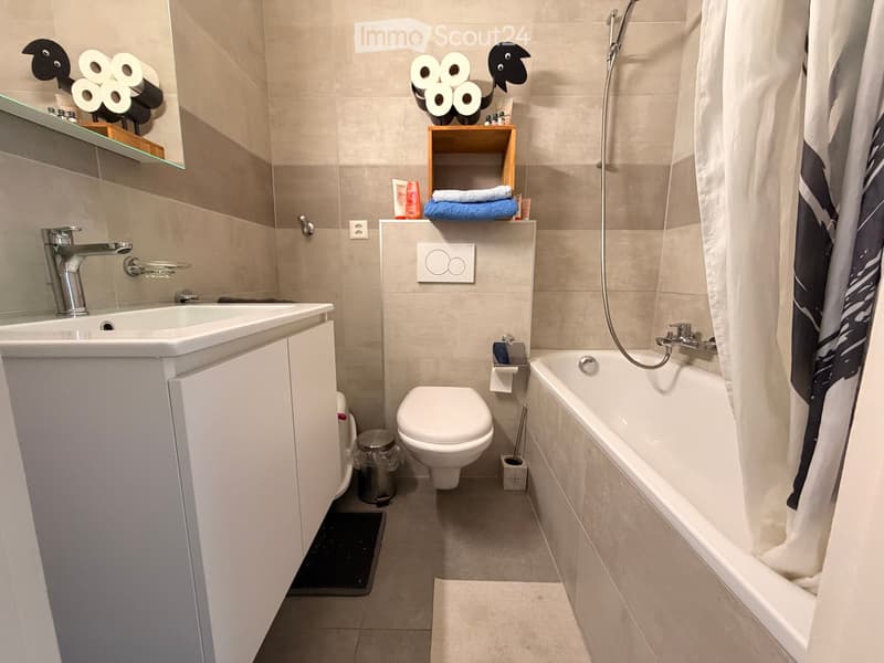 Modern bathroom with a white sink, toilet, bathtub, and a small wooden shelf holding towels and toiletries. The space features beige tiles and a sleek, contemporary design.
