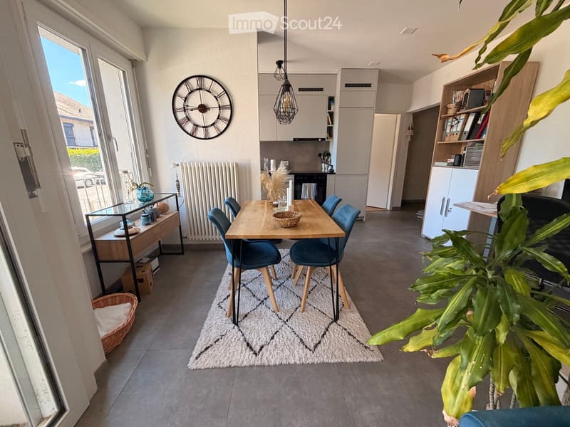 Open-plan living and dining area with a wooden dining table, blue chairs, kitchen cabinets, and a large window offering a view of the outside.