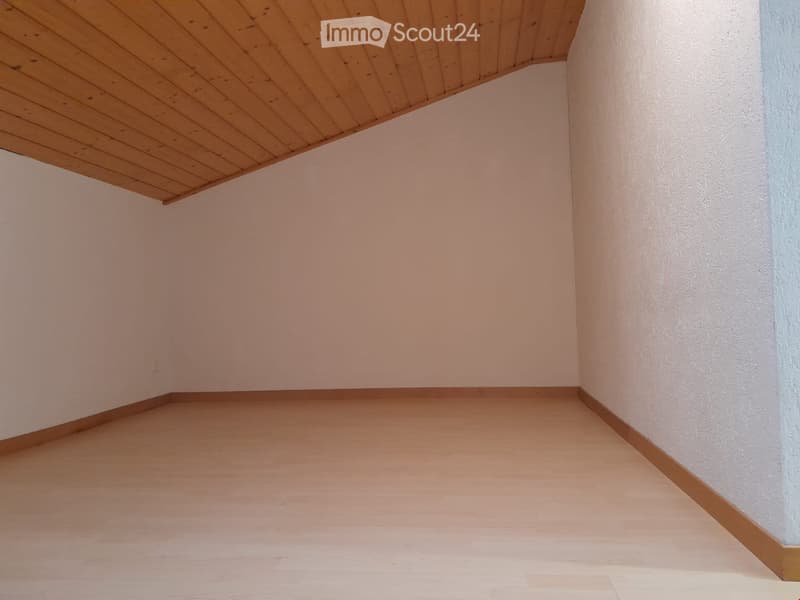 Empty room with light wooden flooring and white walls, featuring a slanted wooden ceiling.