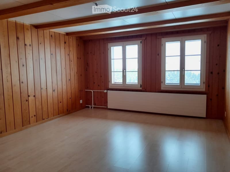 Empty room with wooden walls and floor, large windows, radiator, and exposed wooden beams on the ceiling.