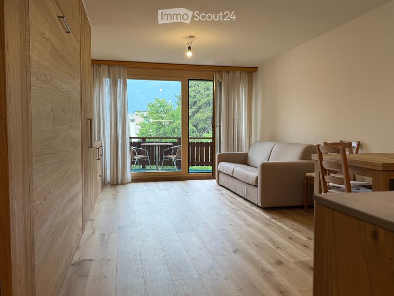 Bright living room with wooden flooring, large windows opening to a balcony, a beige sofa, and a dining area with a table and chairs.