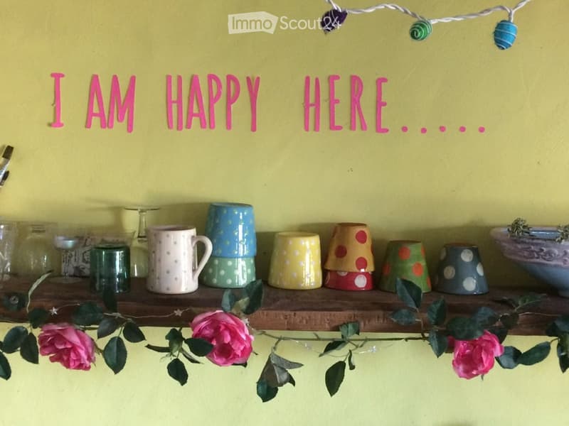 A wooden shelf displays colorful cups and mugs, with a string of lights and a decorative sign reading "I AM HAPPY HERE..." above it. Pink flowers are placed on the shelf.