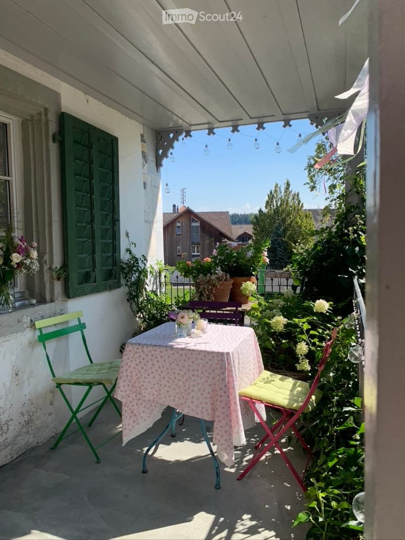 A cozy balcony with a small table and chairs, surrounded by lush greenery and flowers, with a view of the garden and neighboring houses.