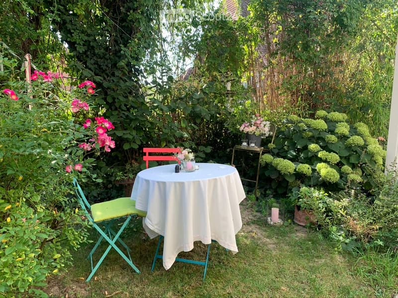 A small, lush garden with a white table and chairs, surrounded by greenery and flowers.