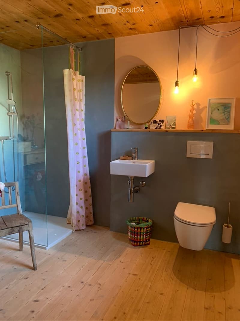 Modern bathroom with wooden flooring, a white sink, toilet, round mirror, glass shower enclosure with a floral curtain, a wooden chair, and a small basket on the floor.