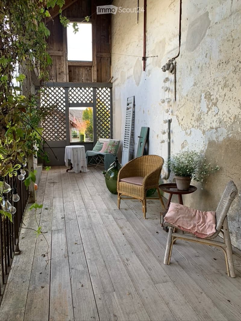 Wooden balcony with wicker chairs, small table, and potted plants, featuring a rustic wooden fence and lattice screen.