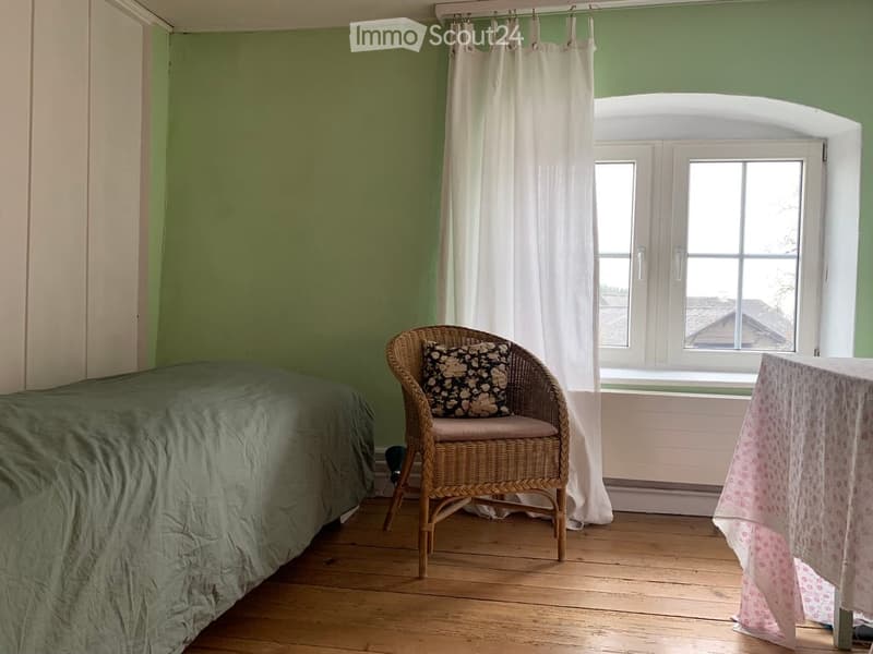 Cozy bedroom with green walls, wooden floor, bed with green duvet, wicker chair with patterned pillow, small table with floral tablecloth, and white curtains.