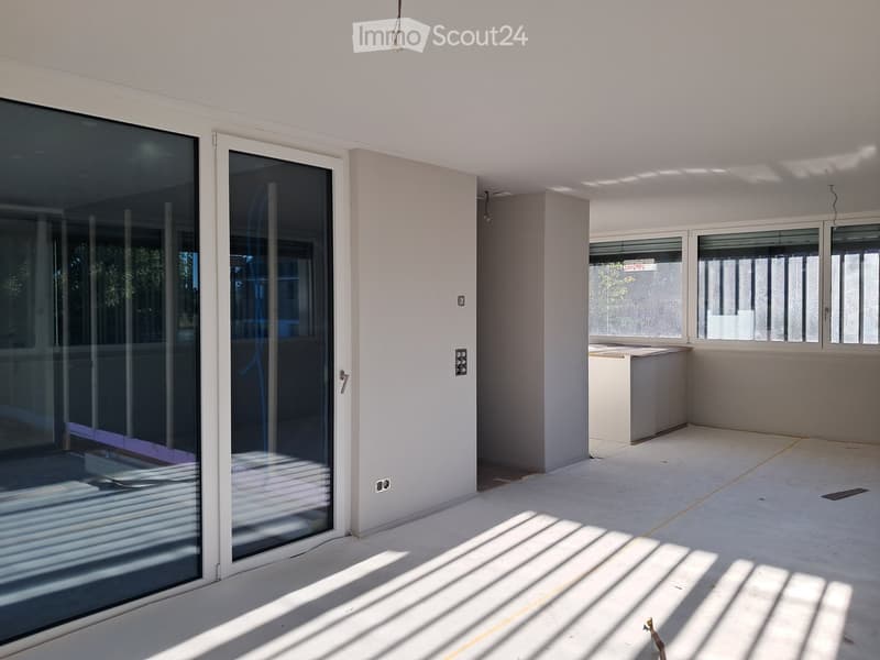 Empty balcony with large glass sliding doors, white walls, and concrete floor. Sunlight streams in, casting shadows on the floor.