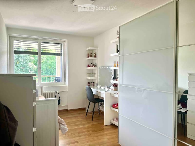 Well-lit room with a vanity area, a large mirror, shelves, a chair, and a wardrobe. The room features wooden flooring and large windows with blinds.