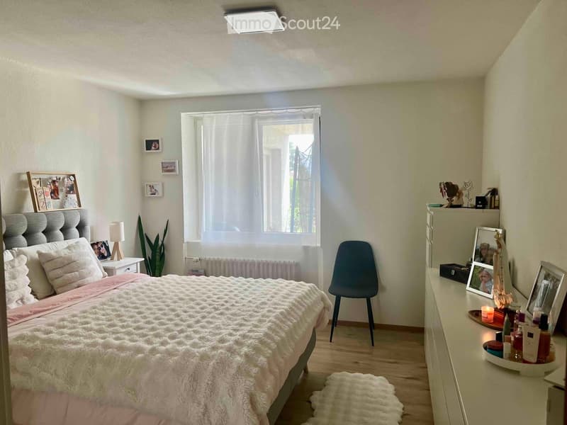 Cozy bedroom with a large bed, nightstand, chair, mirror, and decorative items on a dresser.