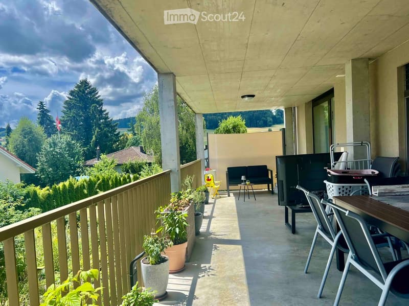 Balcony with a sofa, small table, chairs, potted plants, grill, and storage bin, overlooking a green landscape.