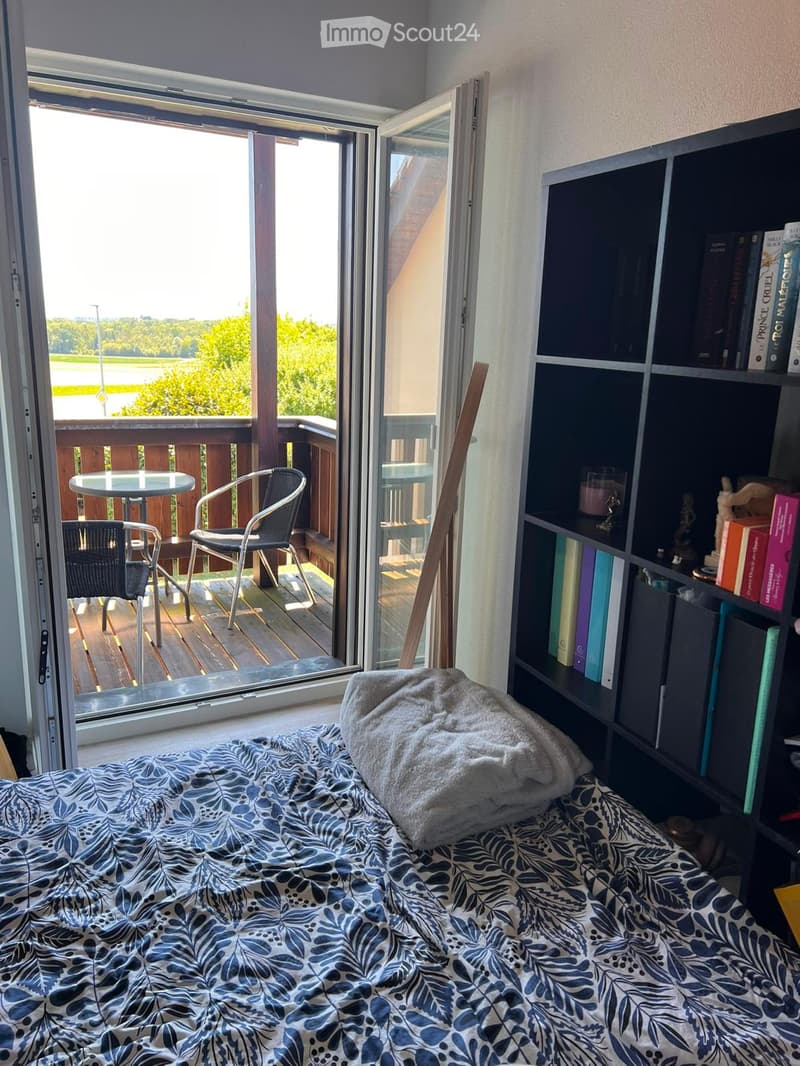 Cozy bedroom with a bed, bookshelf, and a sliding glass door leading to a balcony with outdoor furniture and scenic view.