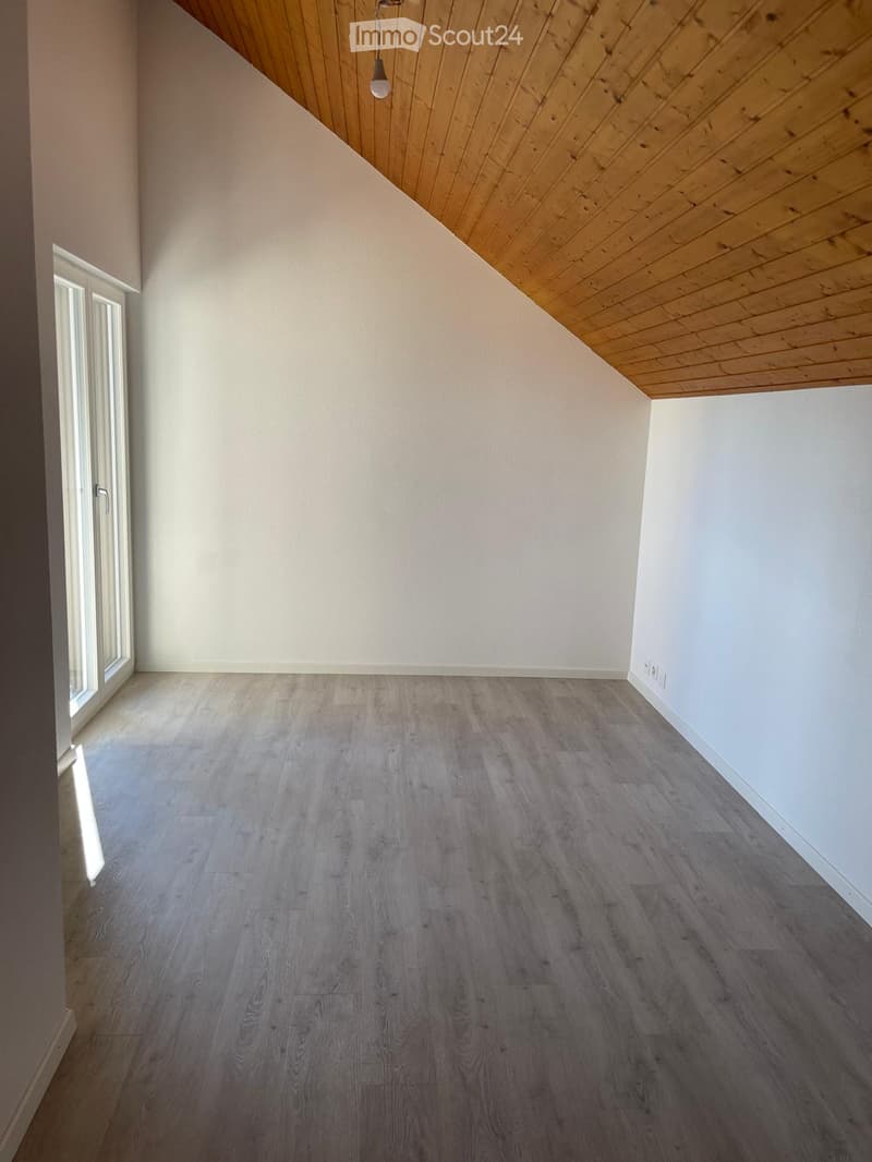 Empty room with light grey wooden flooring, white walls, and a slanted wooden ceiling. A single light fixture hangs from the ceiling, and a large window with white frames is visible on the left side.