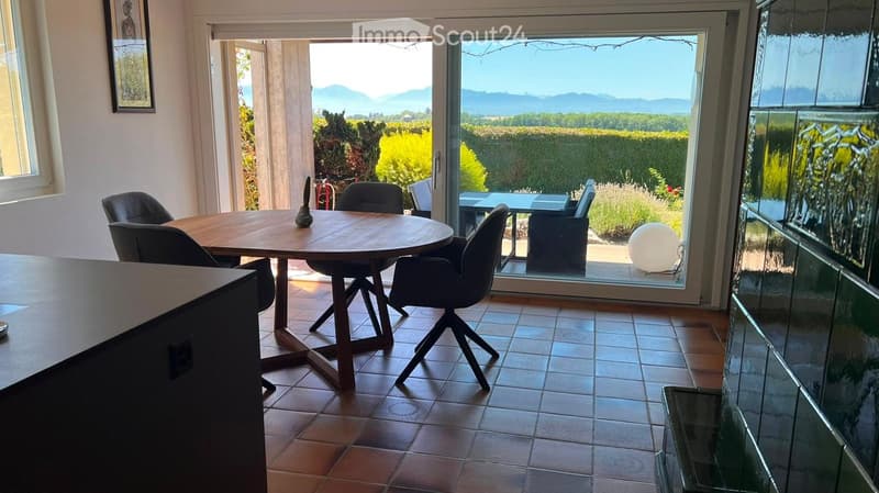 Dining area with a round wooden table, four chairs, and a decorative ball on the table. Large windows offer a scenic view of the countryside.