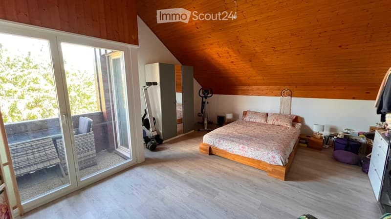 Cozy attic bedroom with wooden slanted ceilings, large glass doors leading to a balcony, a bed with a patterned duvet, a nightstand with a lamp, a mirror, a wardrobe, and an exercise bike in the corner.