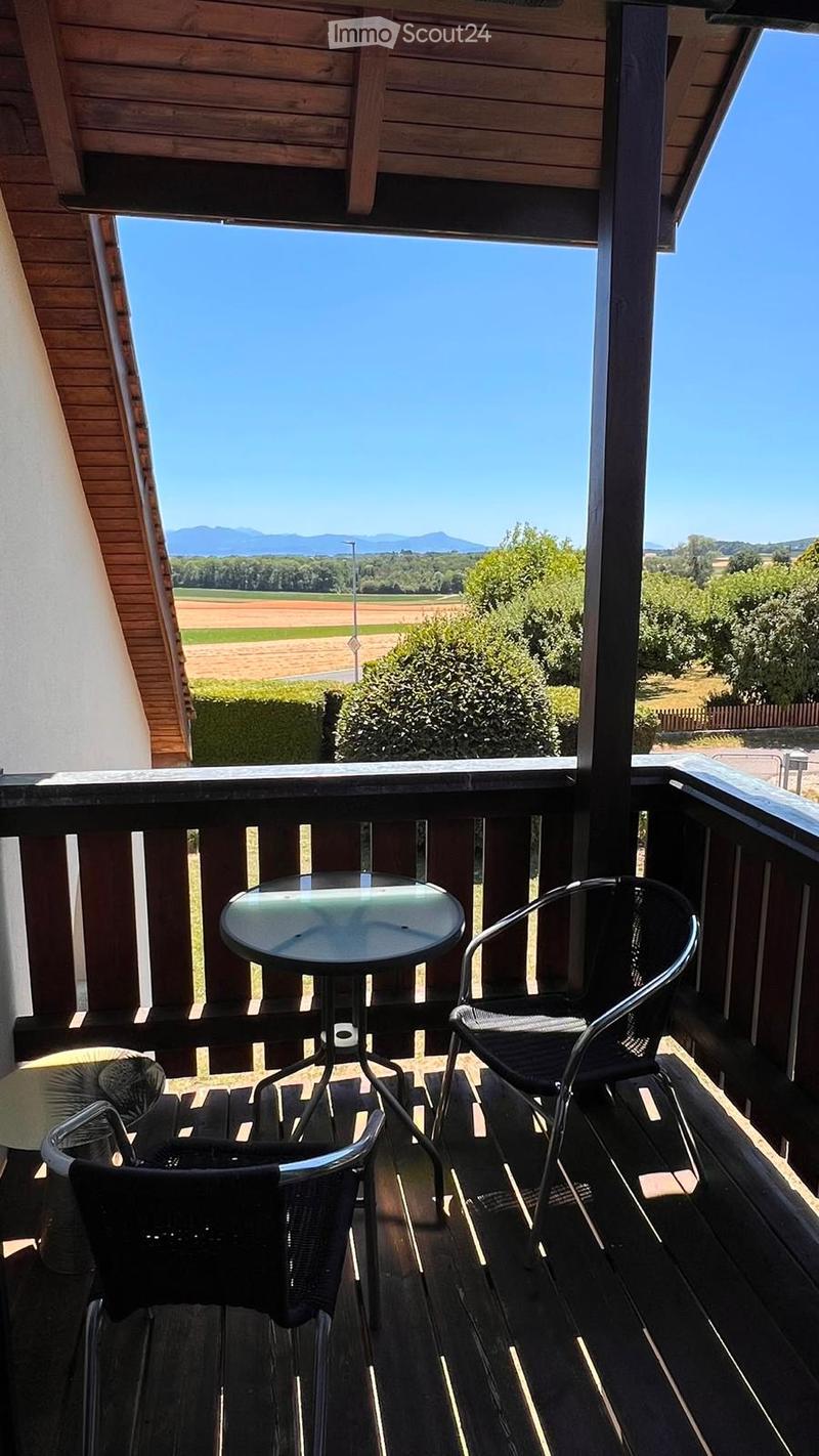 Balcony with wooden decking, a small round table, and two black chairs, offering a scenic view of a countryside landscape with fields and mountains.