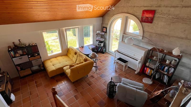 A cozy living room with a yellow sectional sofa, a grey armchair, a white piano, a bookshelf, and a floor lamp. The room features exposed wooden beams on the ceiling and large windows with a view of greenery outside.